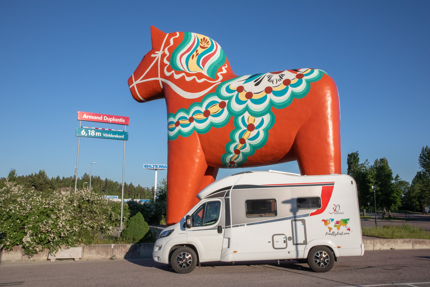En husbil står framför den stora dalahästen i Avesta. Bredvid dalahästen finns en skylt med stavhopparen Armand Duplantis världsrekordhöjd, 6,18. Den höjden går upp till ungefär halva dalahästen.