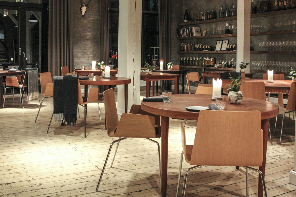 Den danska restaurangen Domestic i Århus. Restauranglokal med runda träbord och karmstolar i trä. Rustikt ljust trägolv och gråa tegelväggar. Tunga gardiner vid sidan om höga fönster.