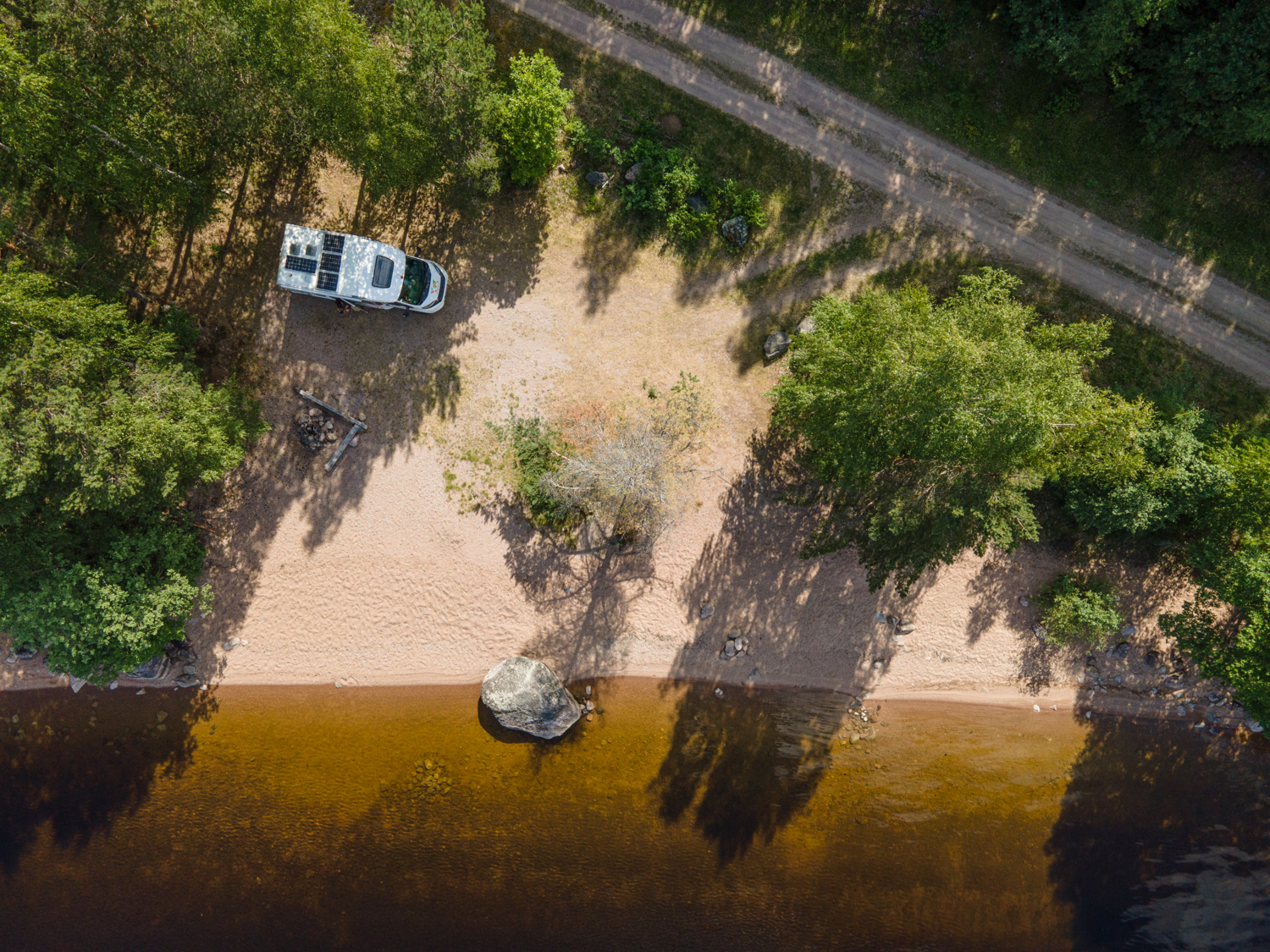 En bild tagen av en drönare. Bilden föreställer en husbil på en sjöstrand vid en skog. Bredvid syns en liten skogsväg.