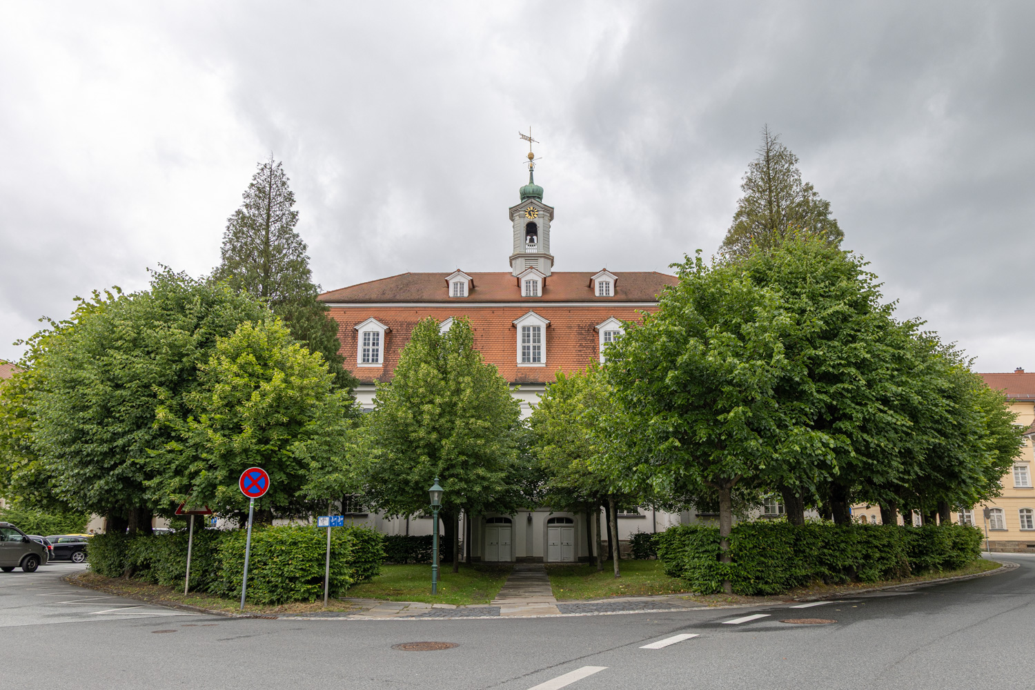 Kyrka i Herrnhut. Ser mer ut som ett stort hus än en kyrka men har ett torn. Framför kyrkan finns stora träd och en häck. Det går en asfalterad väg utanför kyrkans högra sida.
