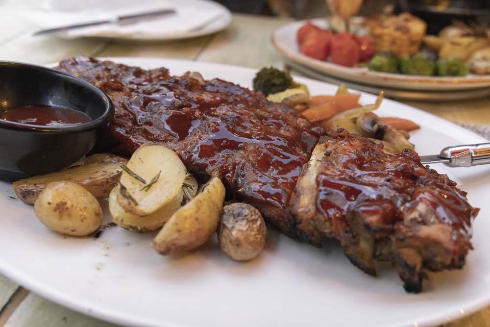 På ett bord syns vit tallrik med revbensspjäll, rostad potatis, sås i skål samt grönsaker.