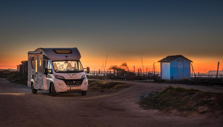 En husbil står vid en strand på kvällen och lyser upp omgivningen. Bakom ser man havet och en solnedgång i vackra färger.