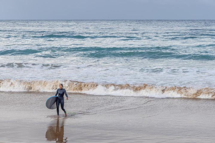 Kvinnlig surfare går ur vattnet med surfbrädan under armen. Bär hellång våtdräkt.