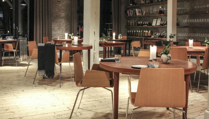 Den danska restaurangen Domestic i Århus. Restauranglokal med runda träbord och karmstolar i trä. Rustikt ljust trägolv och gråa tegelväggar. Tunga gardiner vid sidan om höga fönster.
