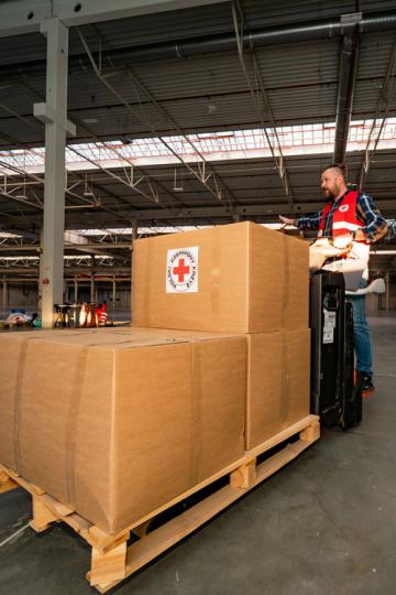 En man på en truck som kör en pall med kartonger från Röda Korset i en nästan tom fabrikslokal.
