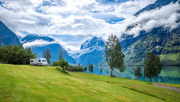 En husvagn står på en gräsmatta med några träd och berg bakom sig. Till höger i bild anas en väg och en sjö.