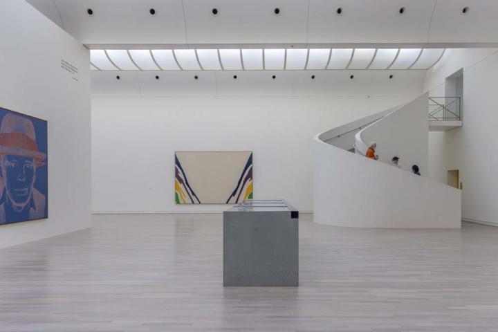 Stort vitt rum med två stora konstverk på väggarna och en monter i mitten. Längst till höger i bild finns en trappa med tre personer som går ner. Annars är rummet tomt.