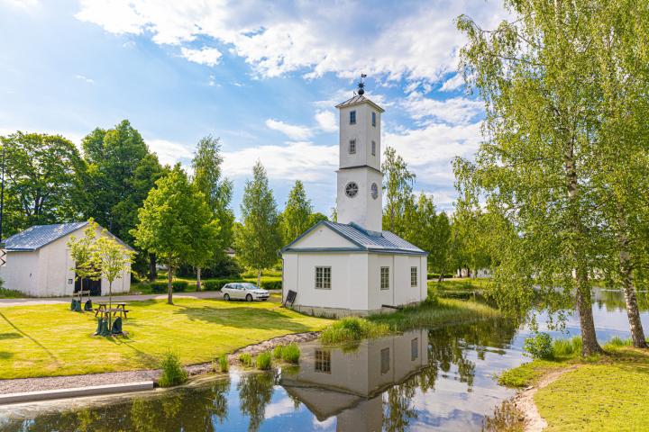 En vit liten kyrka med klocktorn står vid en smal å i ett lummigt landskap.